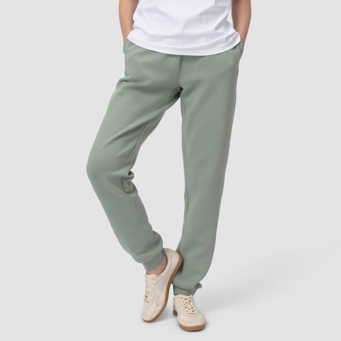 Une personne en tenue de jogging verte, avec des chaussures et un t-shirt blancs, les mains dans les poches