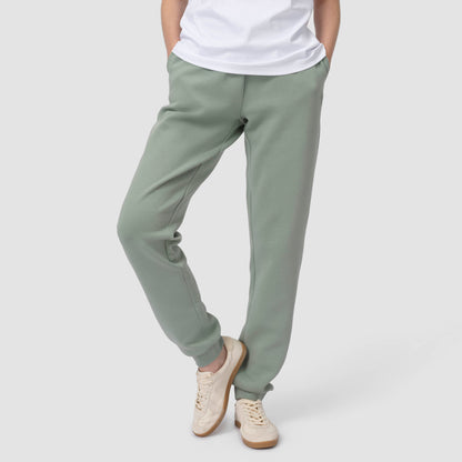 Une personne en tenue de jogging verte, avec des chaussures et un t-shirt blancs, les mains dans les poches