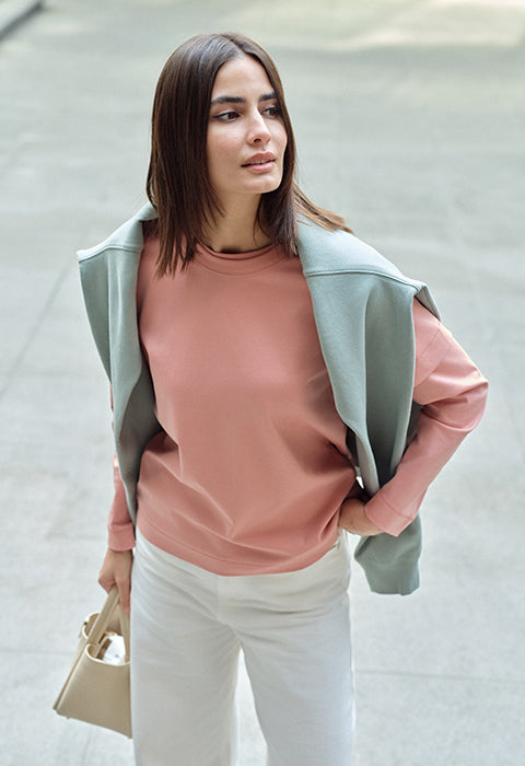 Woman wearing a pink sweater and white pants with a green hoodie, standing on a street.