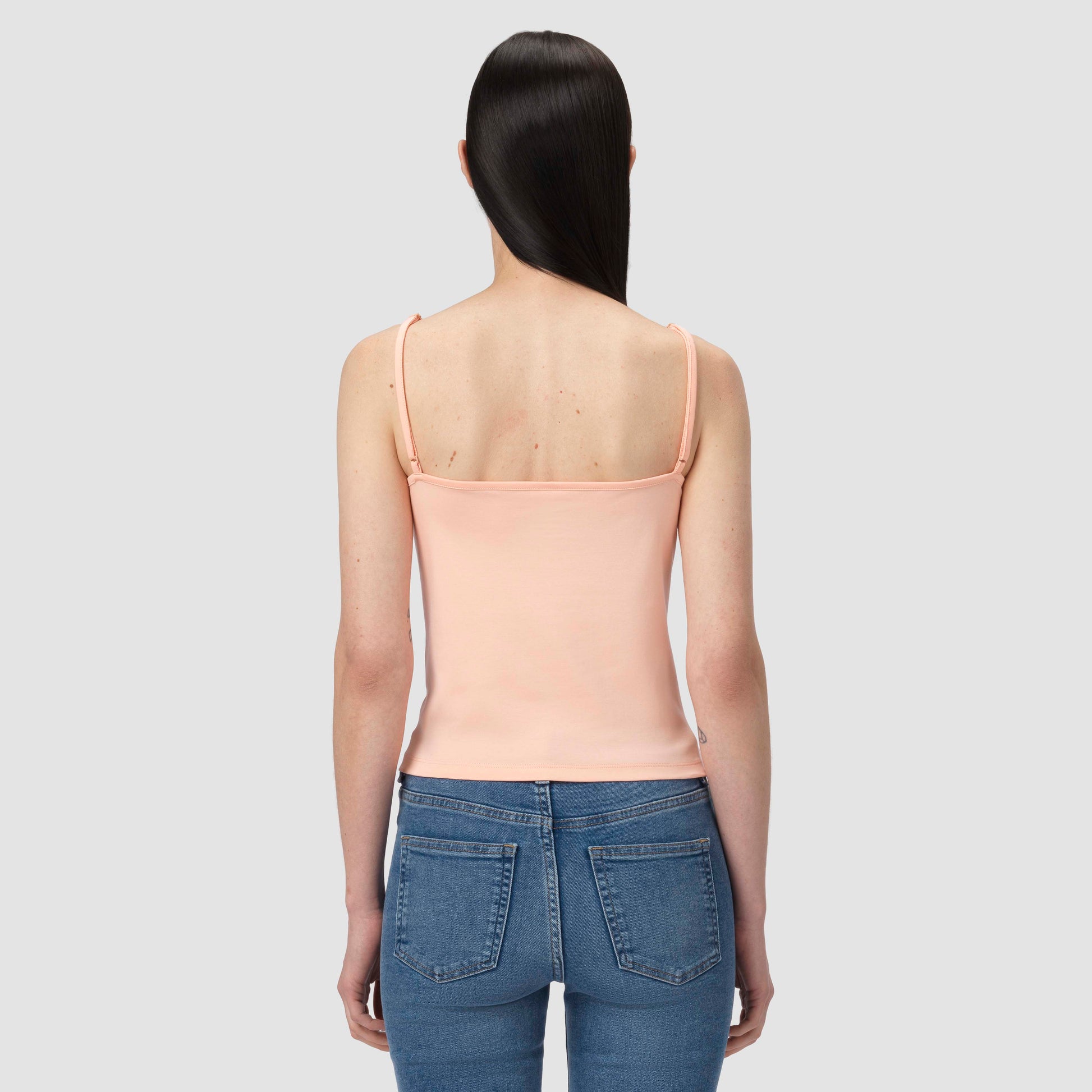 Person wearing a salmon tank top and blue jeans on a light gray background