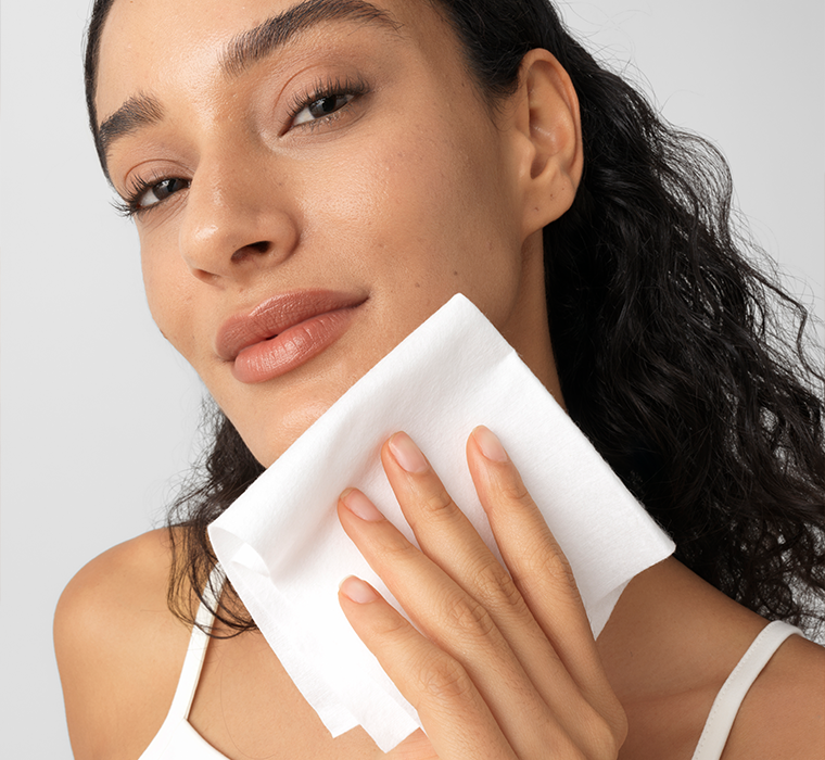 Woman cleaning her face with a white cotton towelette against a plain background