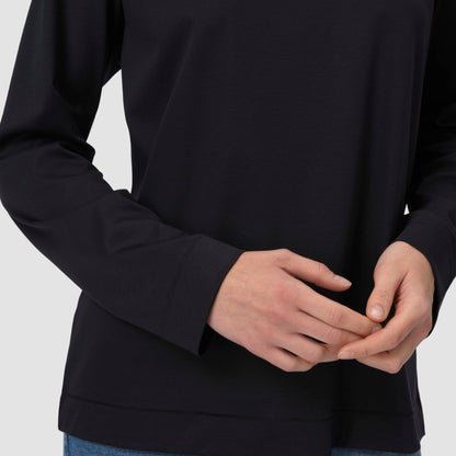 Une personne vêtue d'un t-shirt noir à manches longues, les mains jointes devant elle, sur un fond gris clair